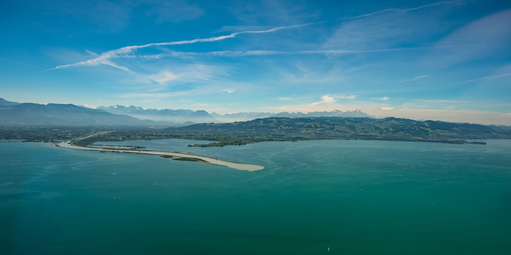 A panoramic view of Lake Constance on a sunny day, with light mist cloaking the surrounding landscape.